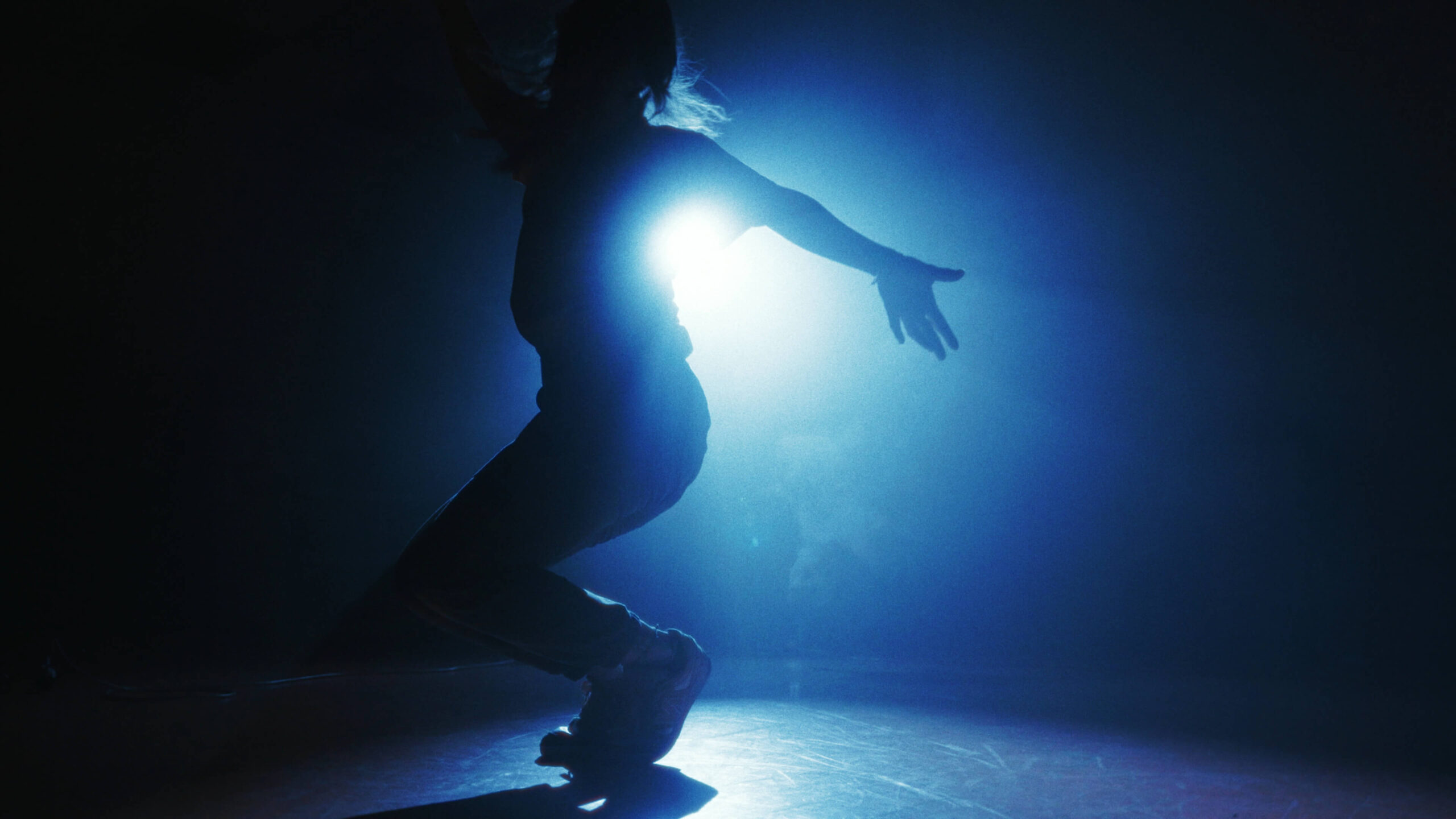 Silhouette de Mahrie Corbu en mouvement de capoeira, éclairée en contre-jour bleu