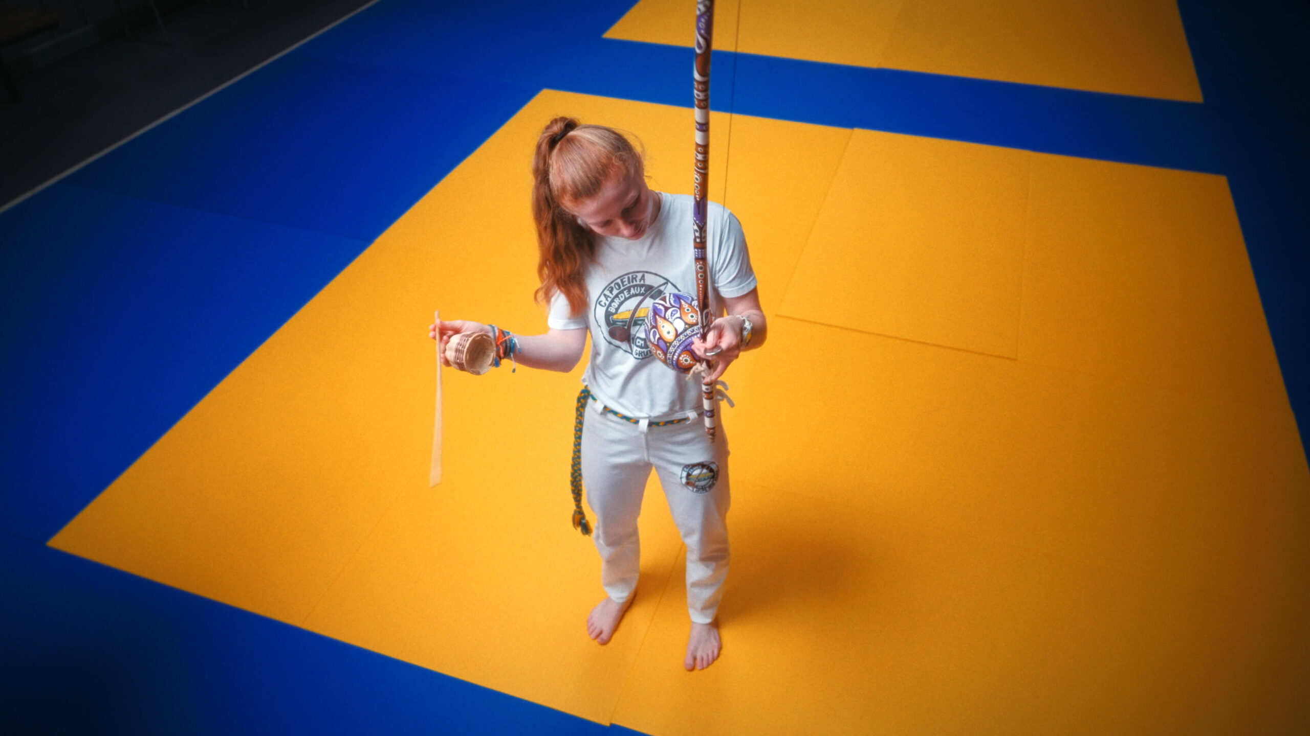 Mahrie Corbu tenant un berimbau et un caxixi sur un tatami bleu et jaune